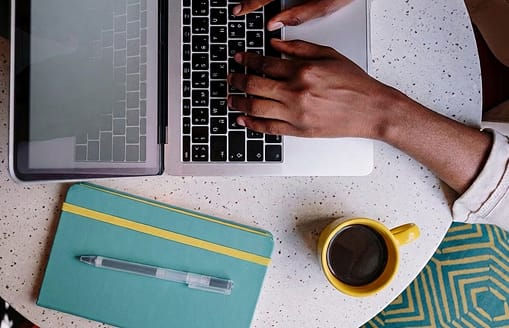 Person on laptop with notebook and cup of coffee next to it on a table